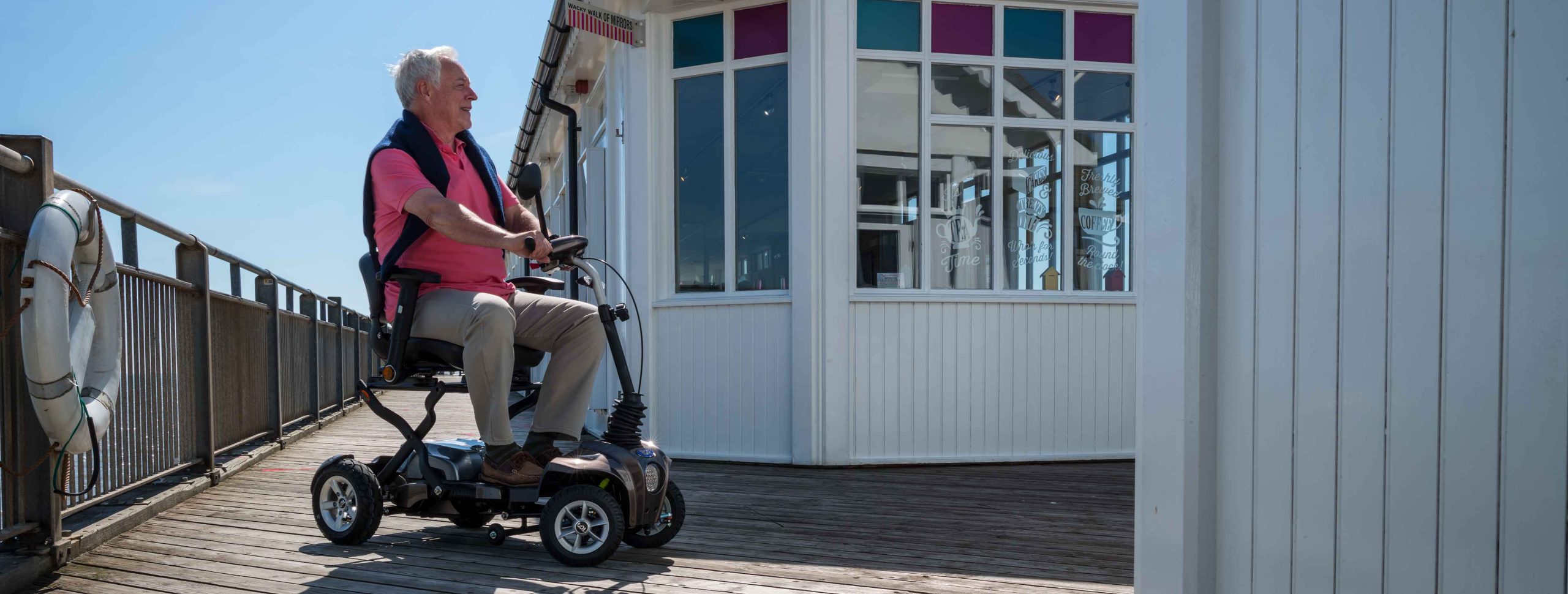 An older man wearing a pink shirt on a folding mobility scooter on a pier